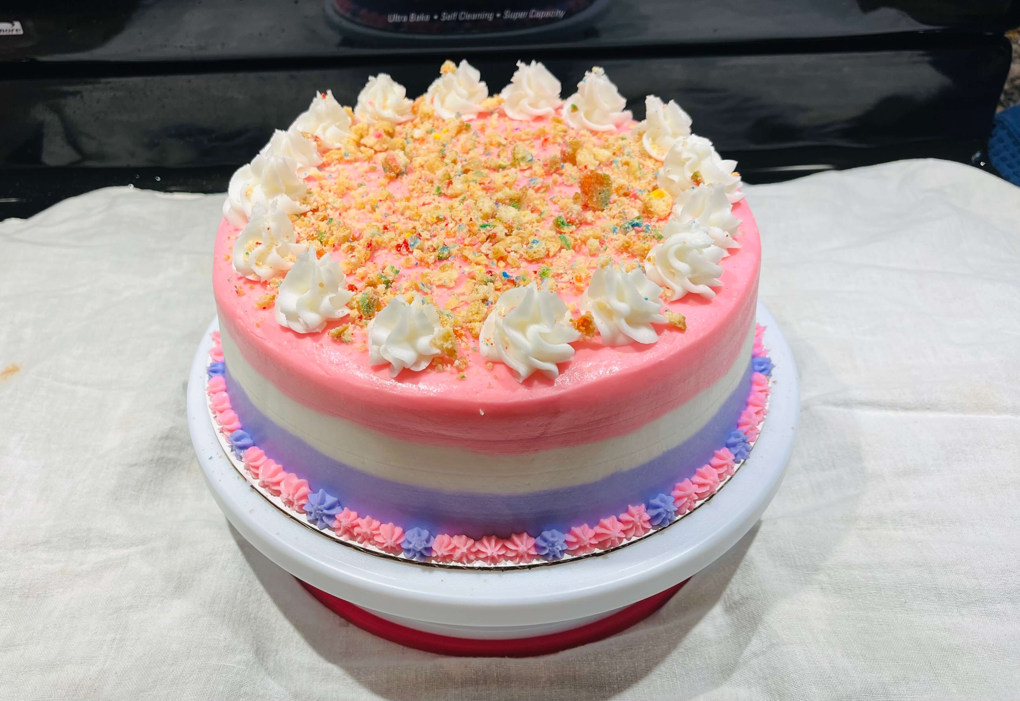 Decorated cake with pink, white and purple bands. Topped with cookie crumbs.